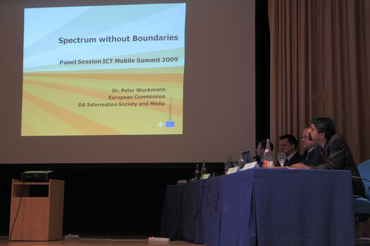 A photo of a panel sitting at a table on a dais. Dominating the frame is a large projection screen displaying a PowerPoint slide with the heading "Spectrum without Boundaries: Panel Session ICT Mobile Summit 2009." Under the screen is a small wood stand with the projector.