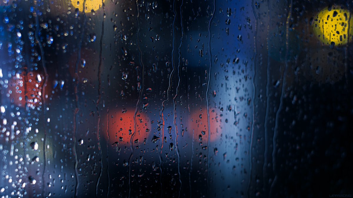 Photography by Liam Wong of Tokyo at night. Images captured on a 50mm lens within a phone booth. This image is the glass with heavy rain drops falling down. The background is soft and out of focus - orbs of colors from taxis and neon signs. It is blue in color with peach and orange highlights.