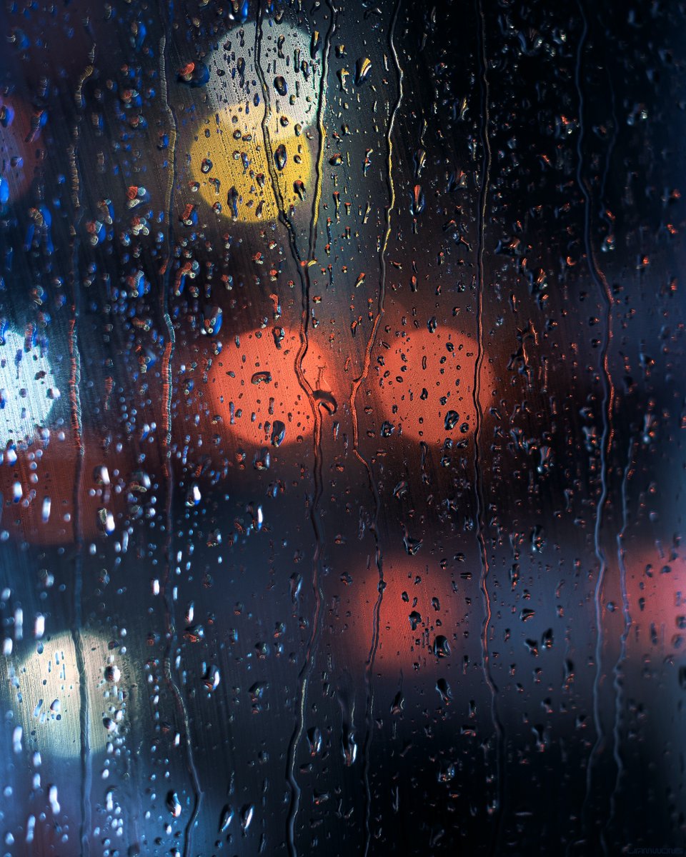 Photography by Liam Wong of Tokyo at night. Images captured on a 50mm lens within a phone booth. This image is the glass with heavy rain drops falling down. The background is soft and out of focus - orbs of colors from taxis and neon signs. It is blue in color with peach and orange highlights.