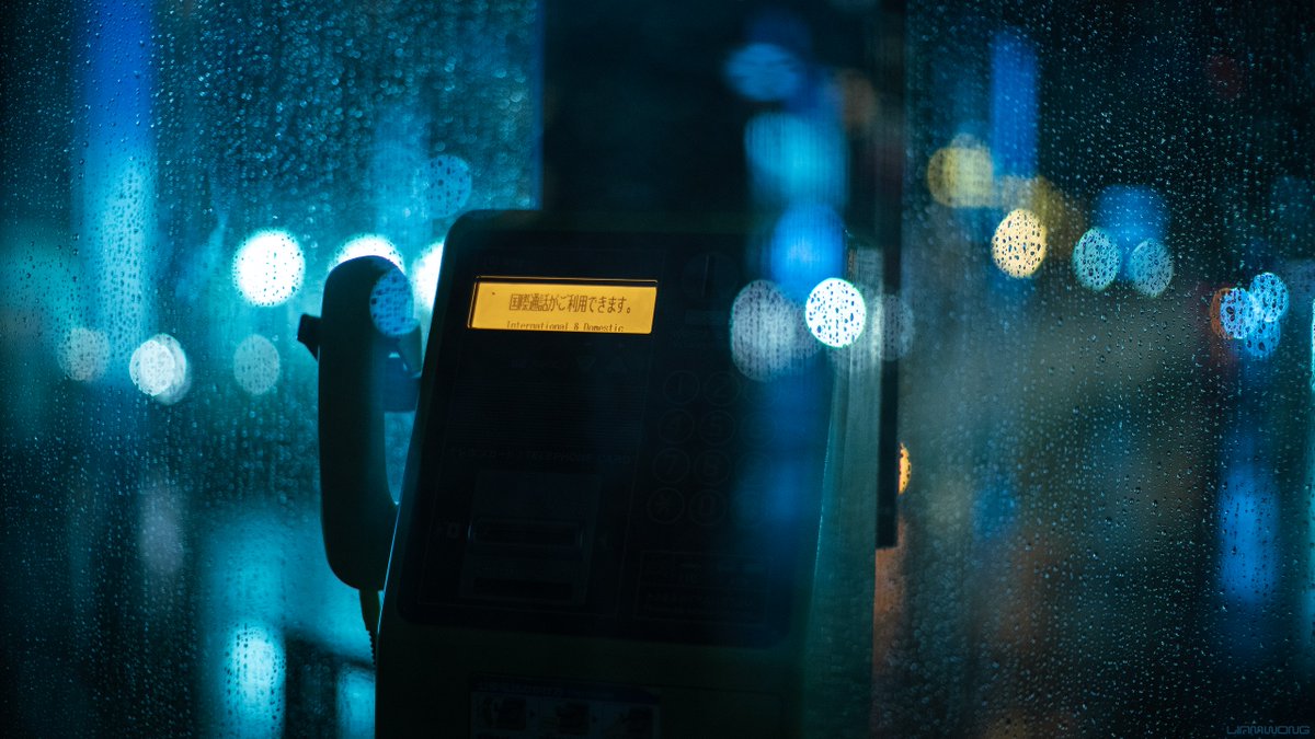 Photography by Liam Wong of Tokyo at night. Images captured on a 50mm lens within a phone booth. This image is the glass with heavy rain drops falling down. There is a phone in focus - it is dark and the light reads International and Domestic (calls) in English and Japanese. It is teal and yellow in color.