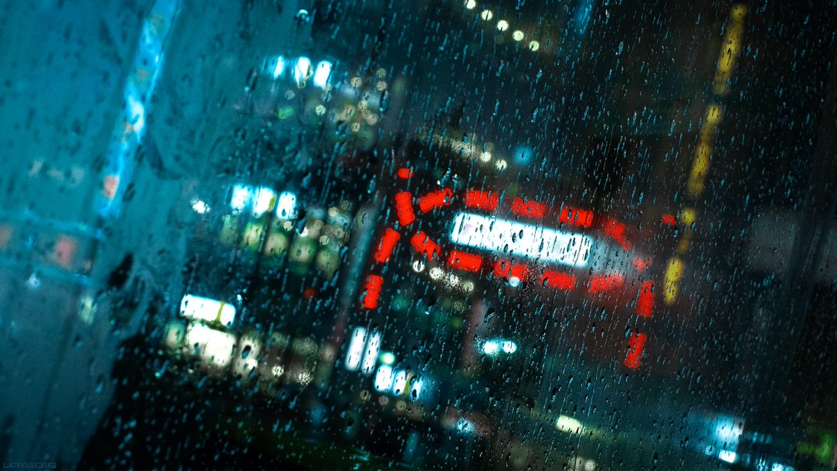 Photography by Liam Wong of Tokyo at night. Images captured on a 50mm lens within a phone booth. This image is the glass with heavy rain drops falling down. The Kabukicho neon sign can be seen blurred in the background. It is teal and red in color.