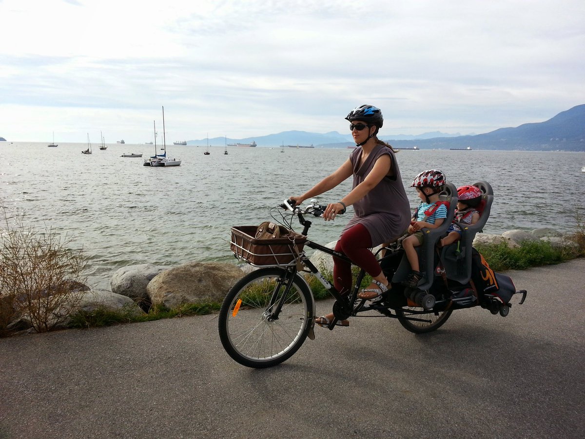 A woman riding a longtail cargo bike with two small kids in seats on the back, a folding stroller strapped to the side, along the sea wall, with sailboats on the ocean in the background