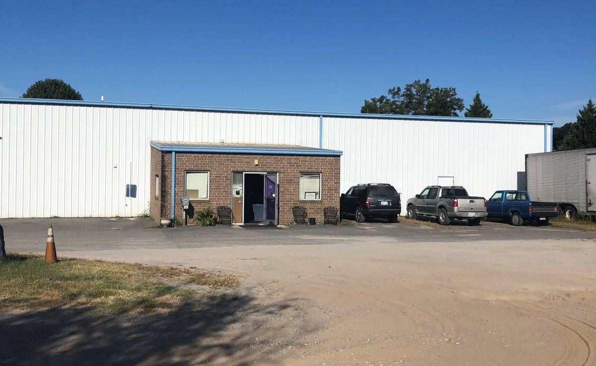 A photo of the small brick entrance to a white vinyl-sided warehouse shot from across a gravel lot. Several trucks are parked outside. There are trees in the background.