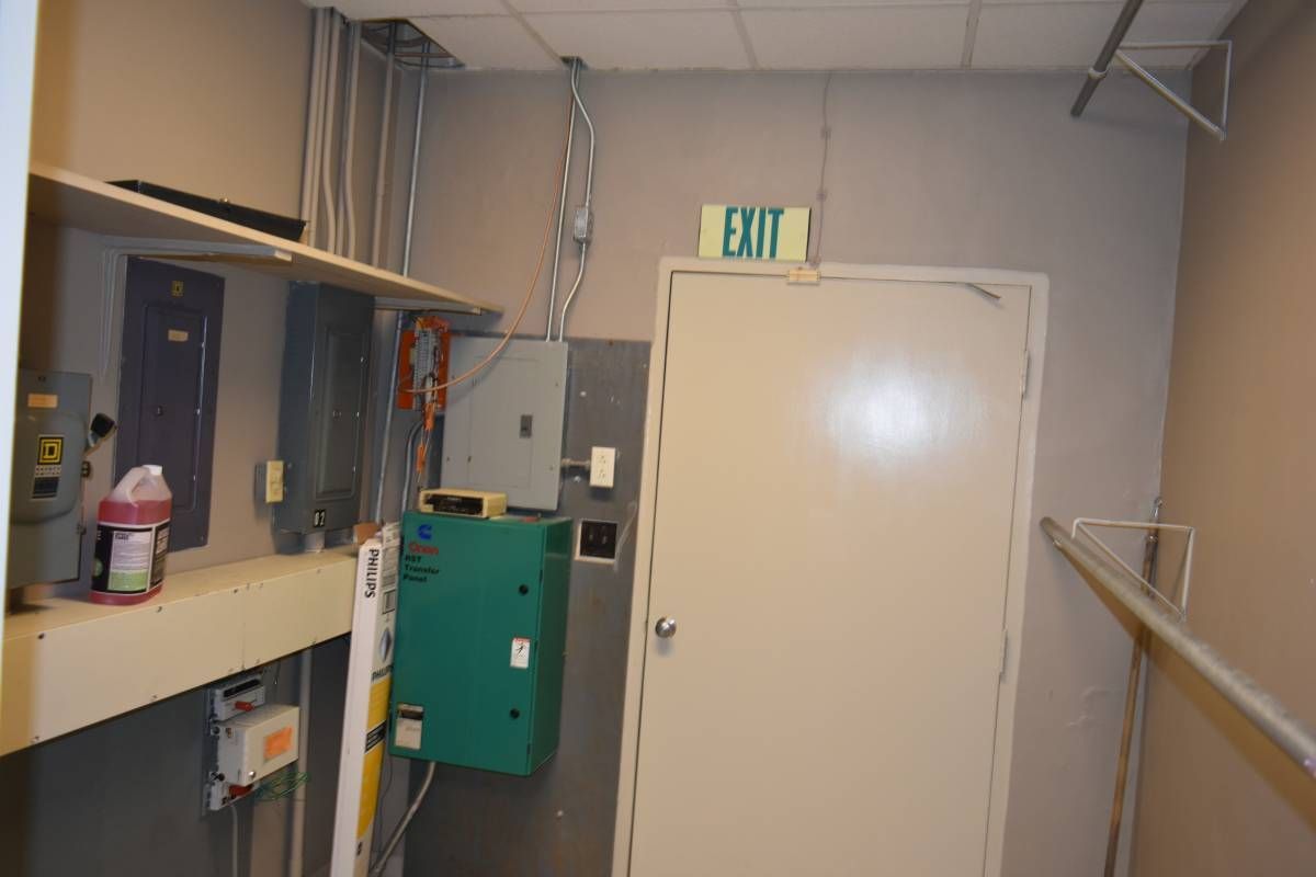 A misaligned flash photo of the back exit door of a commercial building. The door is beige. There are circuit boxes on the wall to the left. There is a jug of cleaning solution on a shelf near an electrical master switch.