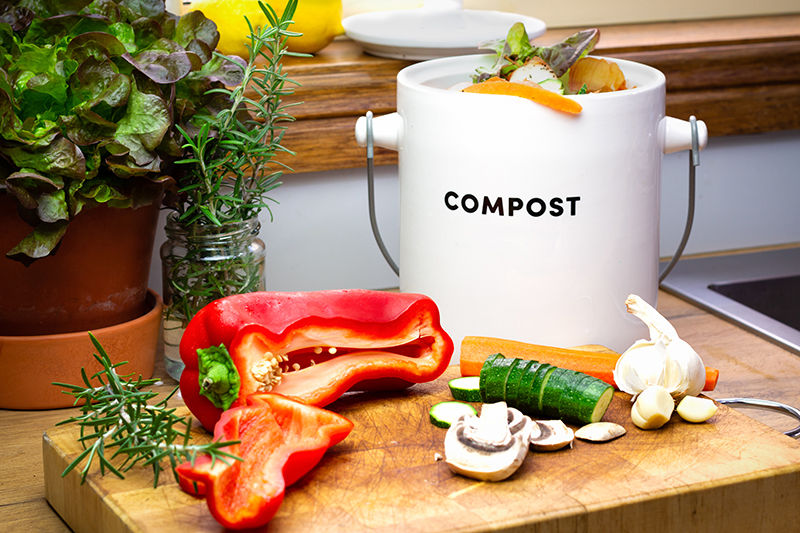 Small white container with the word compost on it, sitting on a cutting board with color vegetables.