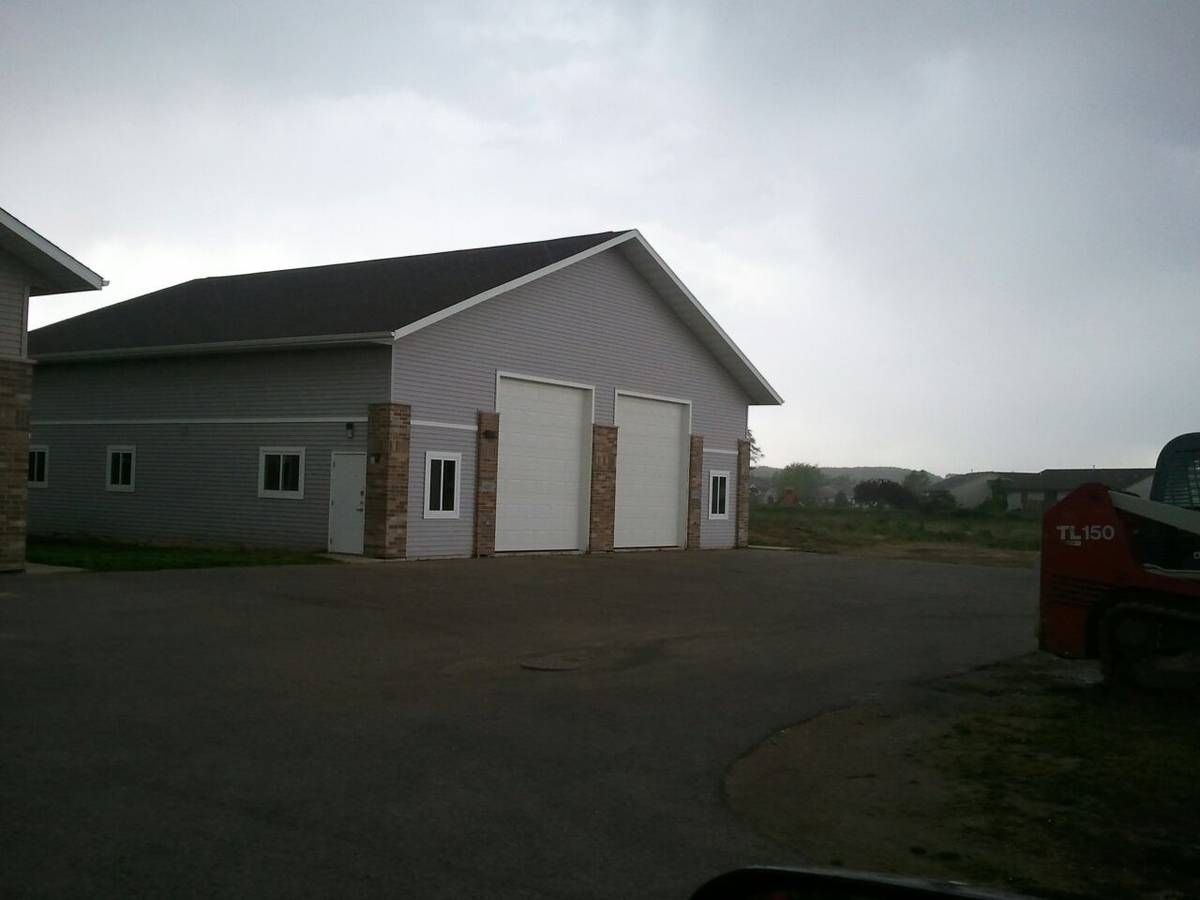 A photo of a gray barn on a blacktop lot. There is a truck parked on the right. There are trees and hills in the distance.