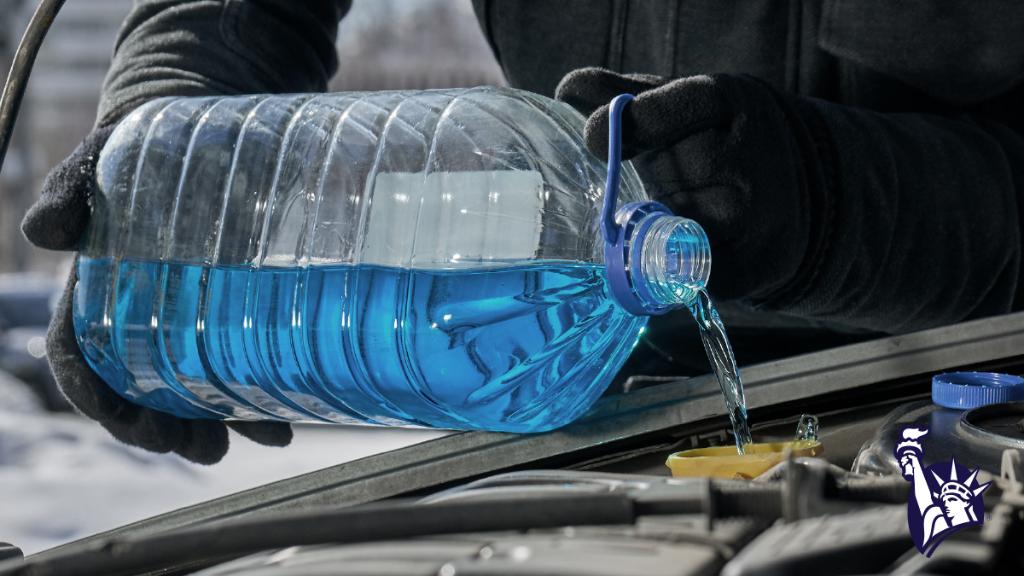 A person wearing a winter coat and gloves pours bright blue windshield wiper fluid into their car.