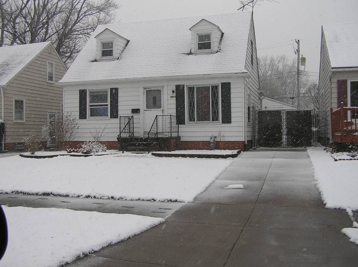 A photo of a white residential house on an old suburban street. Snow is falling and has dusted the rooftops and piled in the yards. In the background, there are trees, a utility pole and power lines.