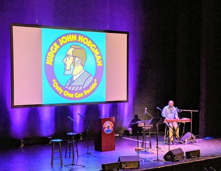 Josh is on stage at The Wilbur Theatre in Boston, seated stage-left and playing organ. The stage is set with stools, microphones, and a podium for a live recording of the Judge John Hodgman podcast. The stage is lit in bright purple. A large screen at the back of the stage displays the Judge John Hodgman logo: a cartoon illustration of Hodgman in profile, adorned with the words, "Only one can decide."