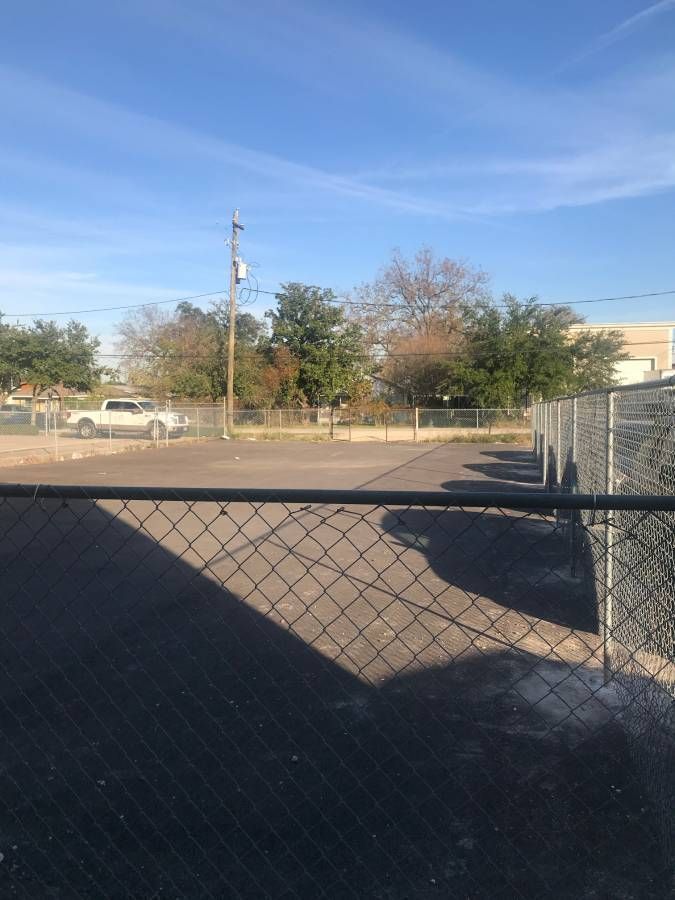 A photo of a parking lot enclosed by a chain link fence. In the background, there are trees, a pickup truck, a utility pole, power lines and buildings.