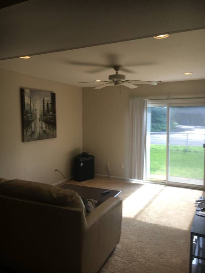 A photo of a room in a residence with beige walls, beige carpet and a beige ceiling fan. A chair faces a sliding glass door standing partway open. There is a view of the back yard enclosed by a fence and a street lined with trees. There is an amplifier in the corner of the room below a canvas with a city image. There is a shelf unit visible on the right.