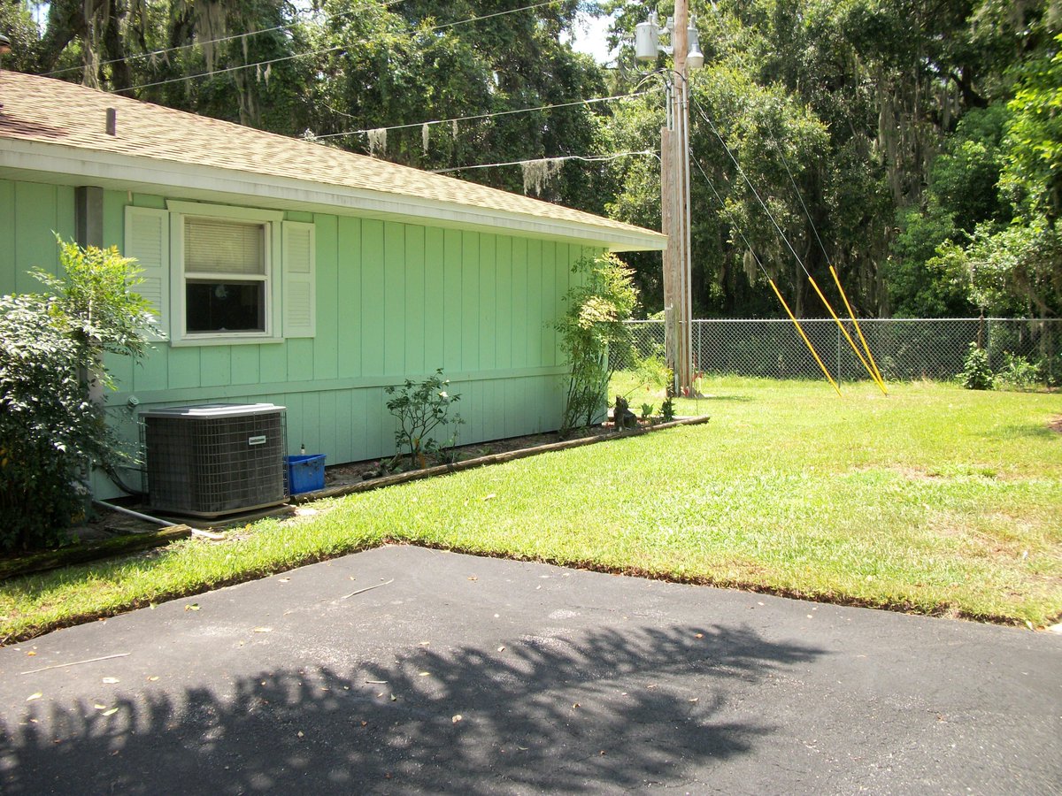 A photo of the fence-enclosed back yard of a suburban ranch-style house. There is a blacktop drive here surrounded by a lawn. Small shrubs are planted in a landscaping strip around the house. There is a utility pole with support cables and power lines. In the background, there are trees.