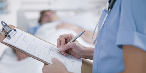 A nurse fills in a chart as a patient (blurred) lays in bed