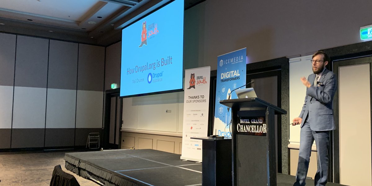 Neil on stage giving a talk at podium with screen showing intro slide and sponsorship banners.