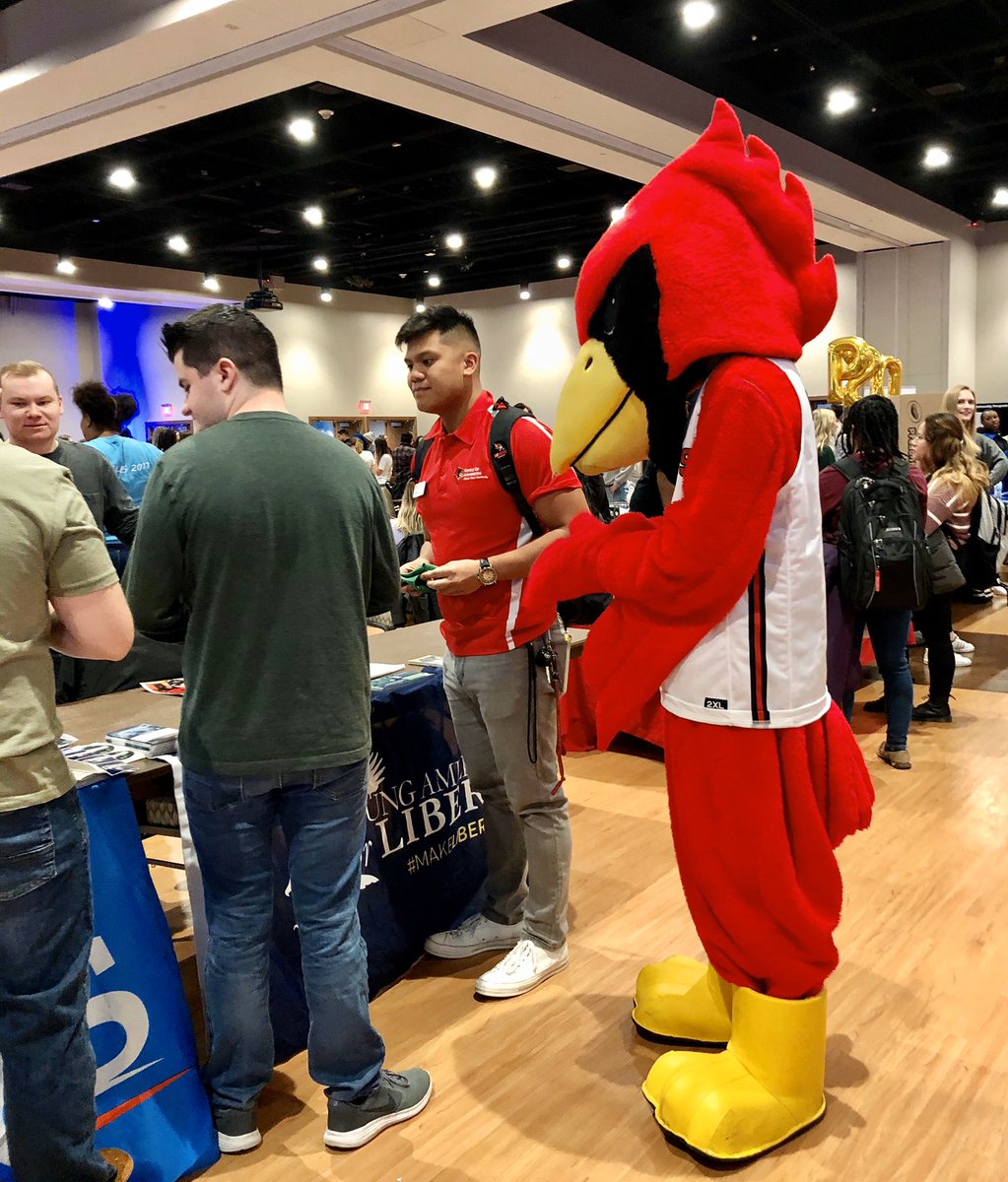 Reggie Redbird interacting with students inside the Brown Ballroom. Floors are beige and shiny with the background exposing the ceiling and lights of the venue.