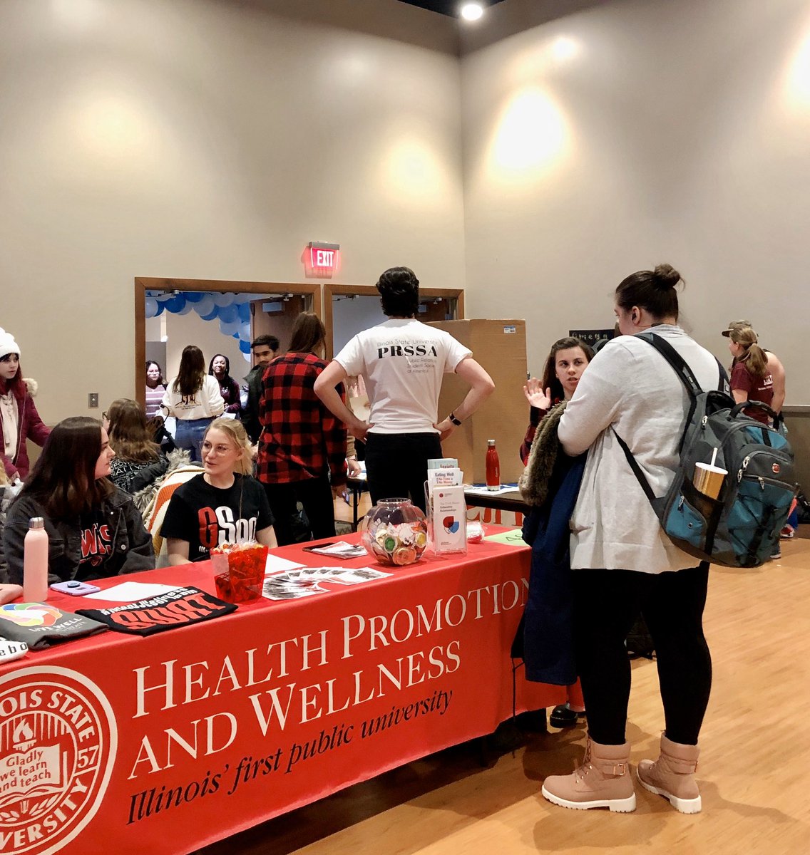 Red table banner for Health Promotion and Wellness has students socializing around towards one of the exits in the room.