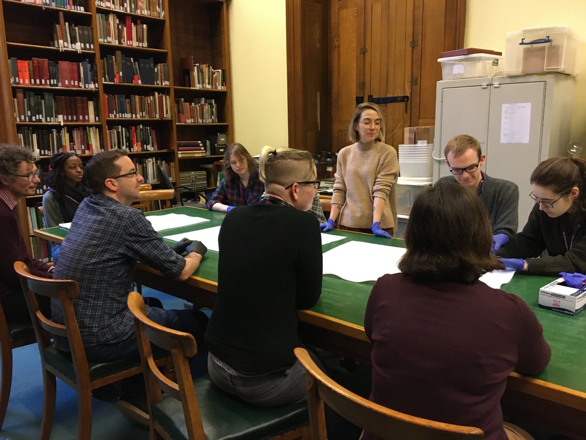 Eight people sit around a table, with one woman standing in the middle. They are all looking at a small object one of the seated people is holding. The room has a bookcase on one wall in the background, and a filing cabinet on the other wall