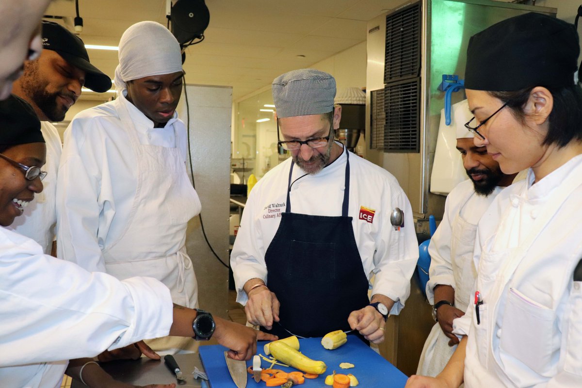 A chef, center, in a chef's coat and apron is demonstrating how to chop yellow squash. 6 participants from the program stand around him watching. One woman in a black hat and glasses excitedly points at the squash.
