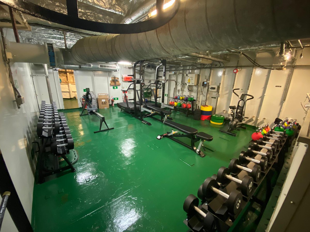A compartment within a ship, white bulkheads, green deck. Lots of sports equipment, weights and exercise machines