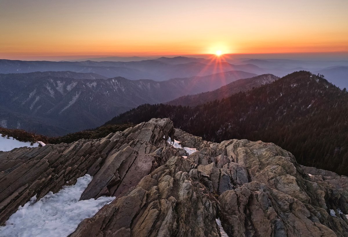 From the summit of a rocky mountain dusted with snow, a view of rolling mountains stretches out to the horizon with an orange sun setting in a clear sky.