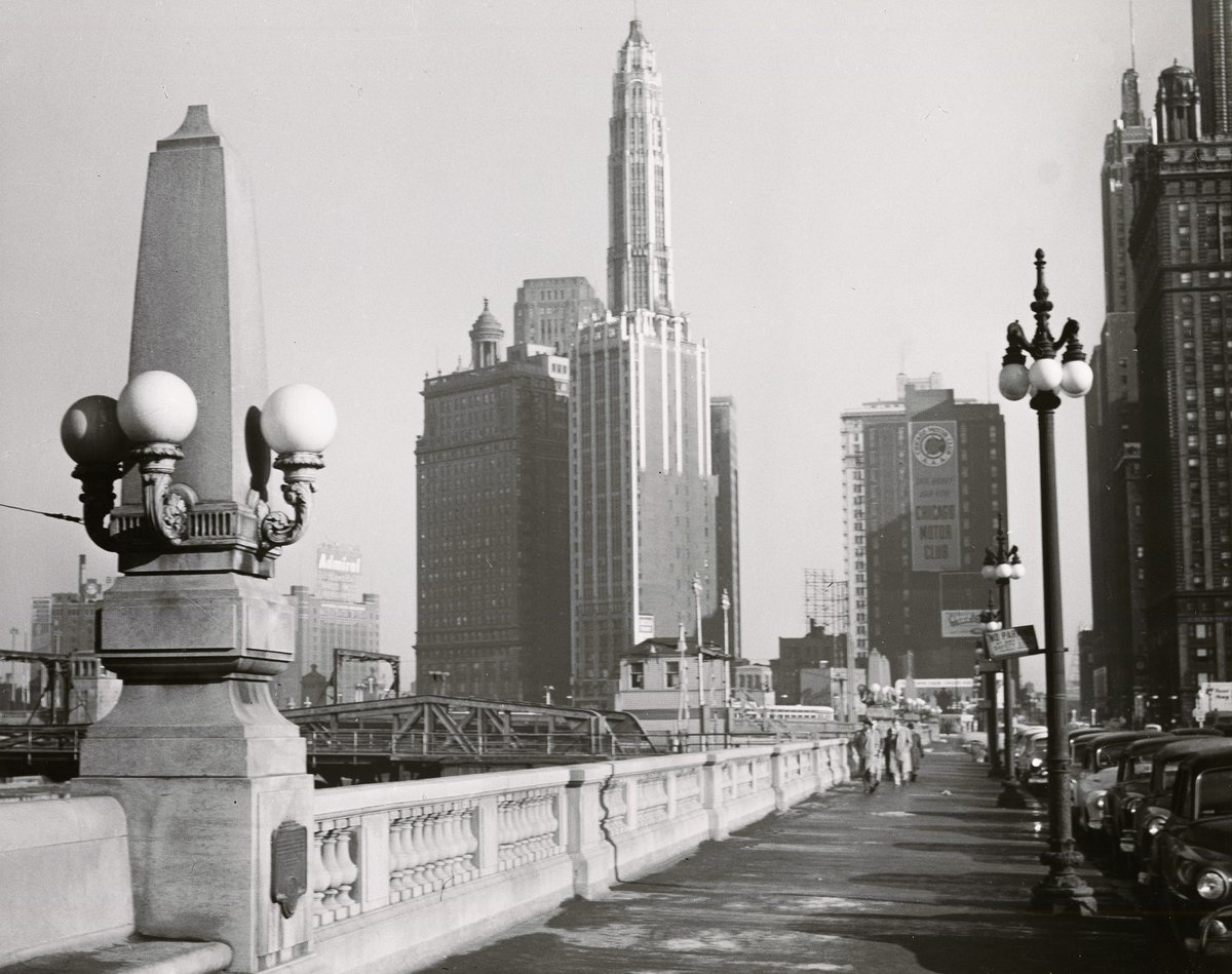 A wide sidewalk with pedestrians along Wacker Drive in downtown Chicago. An unpictured river is on the left, but 2 bridges can be seen in the photo. Streetlights and cars line the road, and there are tall buildings in the distance.
