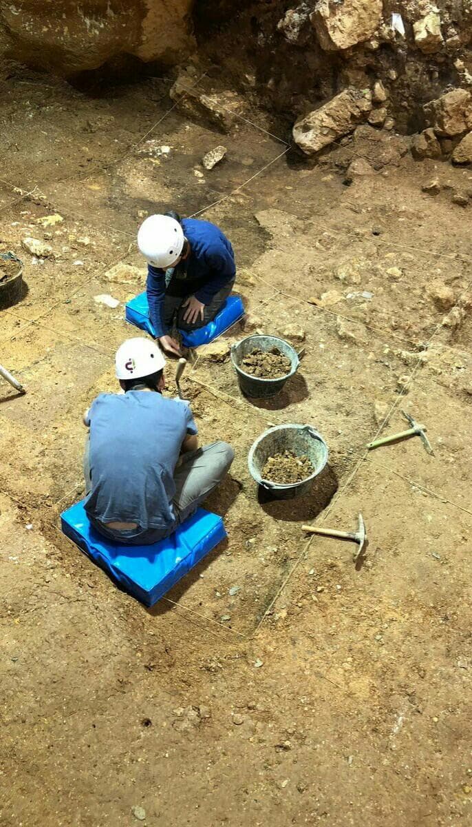 Olalla and another person in a dig site. They are wearing helmets and kneeling on cushions on the ground with rock hammers.