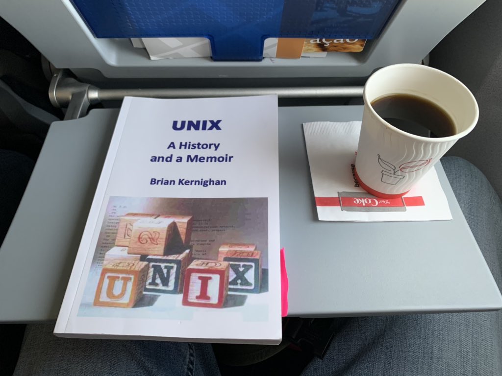 A book, "UNIX: A History and a Memoir" by Brian Kernighan, rests on a airplane-seat tray table next to a cup of cheap coffee.