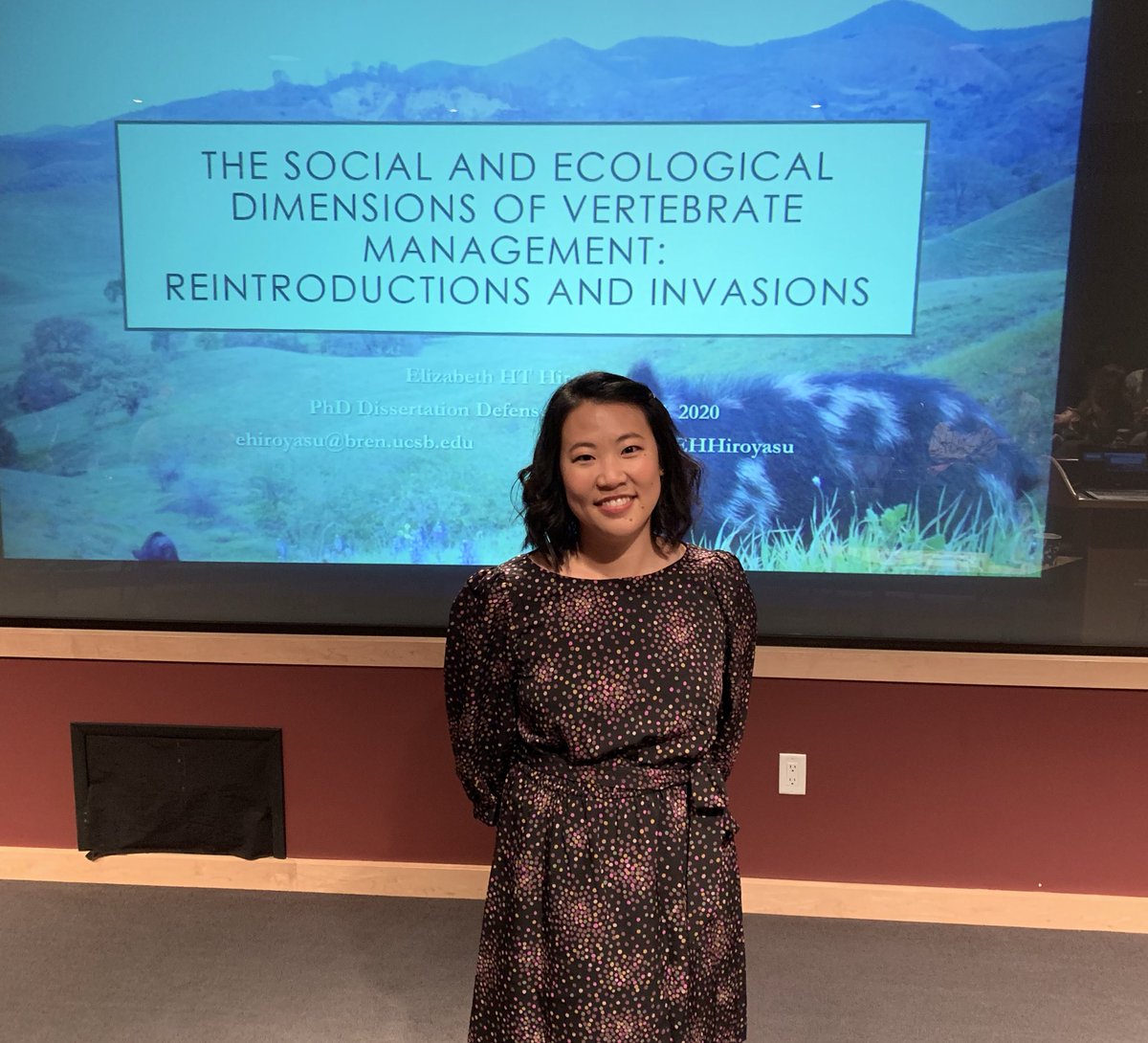 Smiling woman stands in front of projector screen displaying name of presentation.