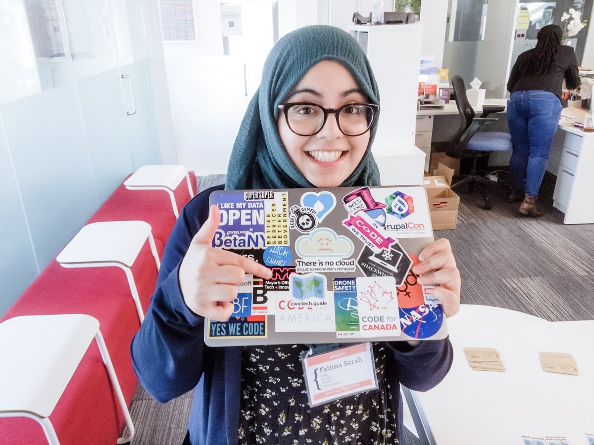 picture of Fatima smiling and holding her laptop of stickers and pointing to the civic tech guide sticker. 