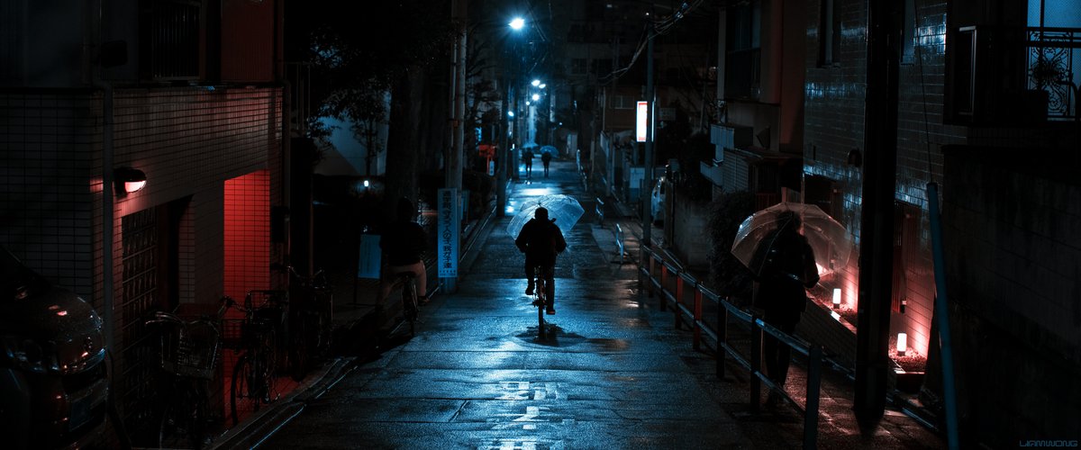 Photography by Liam Wong of Tokyo at night. The silhouette of a man holding an umbrella cycles down the exact centre of the shot. It is blue and red in color. The red light is from an apartment. 