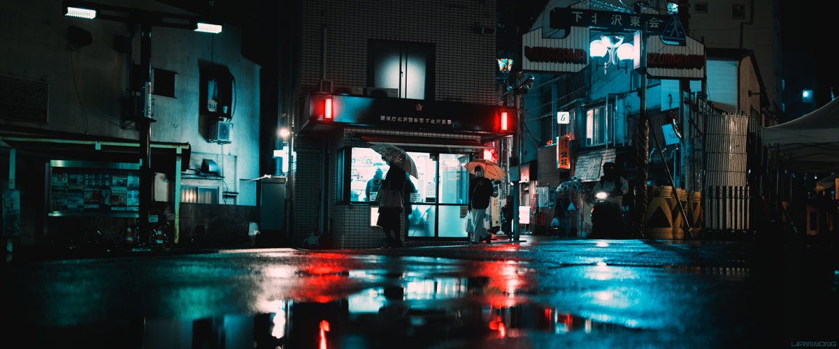 Photography by Liam Wong of Tokyo at night. A woman stands outside a police station checking her phone whilst holding her umbrella in the rain. The red lights from the police station surround her silhouette. It is blue and red in color with most other colors stripped away for dramatic effect.