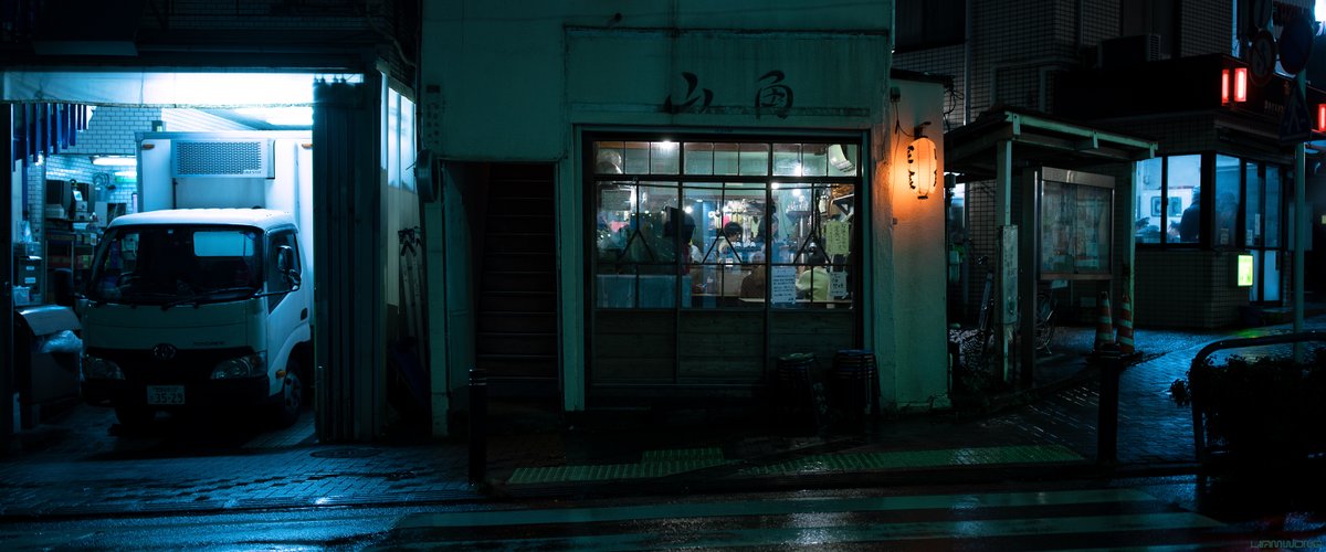 Photography by Liam Wong of Tokyo at night. A picture of a restaurant front-on I the rain. Many people ca be seen inside the window, made up of several panels. A van is lit in a blue light on the left side. On the right side, two policemen are seen in a Konban (police station) speaking to two silhouettes.