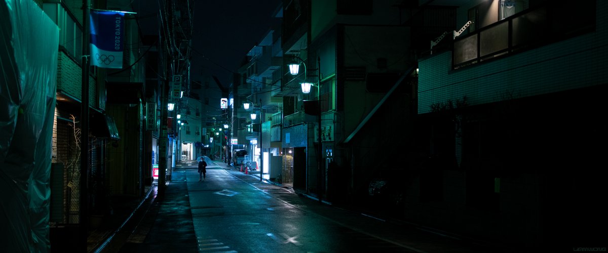 Photography by Liam Wong of Tokyo at night. The silhouette of a man holding an umbrella ca be seen in the distance. The street is eerily quiet. A small banner on a lamp sign says ‘TOKYO 2020’ with the olympics logo made up of four connected rings. It is green in color, with many white lights all the way down the street.