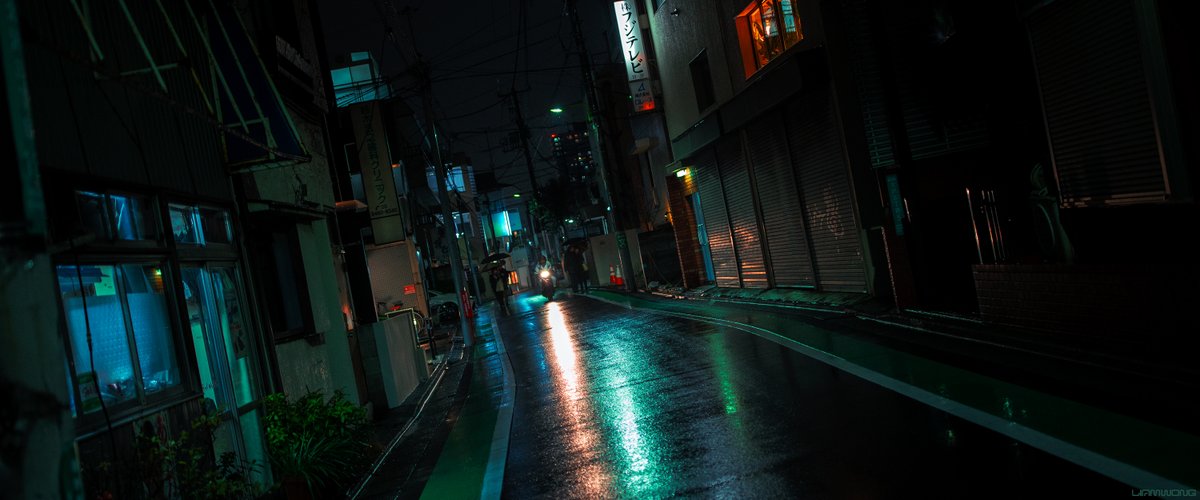 Photography by Liam Wong of Tokyo at night. A ‘dutch angle’ photograph of quiet street in Tokyo in the rain. A motorcyclist is a short distance from the camera, surrounded by people with umbrellas. The light from the motorcycle is lighting up the street. To the left and right are blue and orange colored lights in the widows. It is angled in an obscure way.