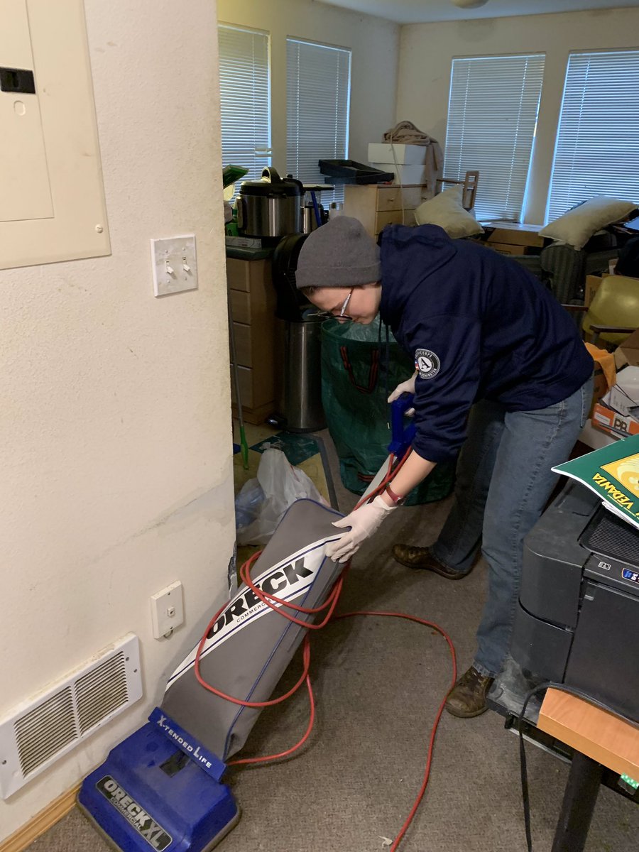 A person wearing a dark blue sweatshirt vacuums grey carpet in an apartment.