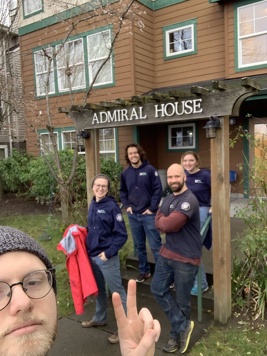 A person takes a selfie of a group of four other AmeriCorps members wearing blue sweatshirts and standing outside a building labeled Admiral House.
