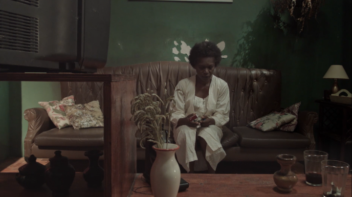 Still from a film to be shown at "Visions of Resistance," featuring a dark-skinned woman sitting on a couch in a dated living room. The room is very dim.