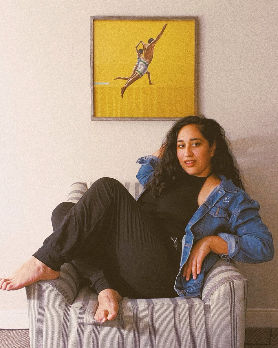Annie sits horizontally across chair under framed yellow artwork, wearing a denim jacket and wavy hair, with one leg tucked under her and the other hanging over the arm of the chair.