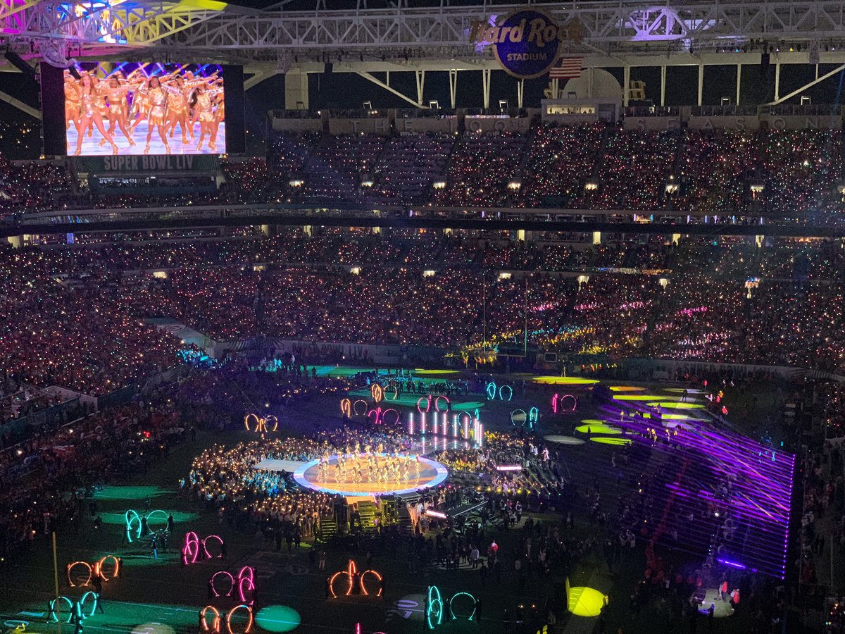Colored lights and light rings shine on the on-field stage at the Super Bowl halftime show. Dancers can be seen on a big screen.