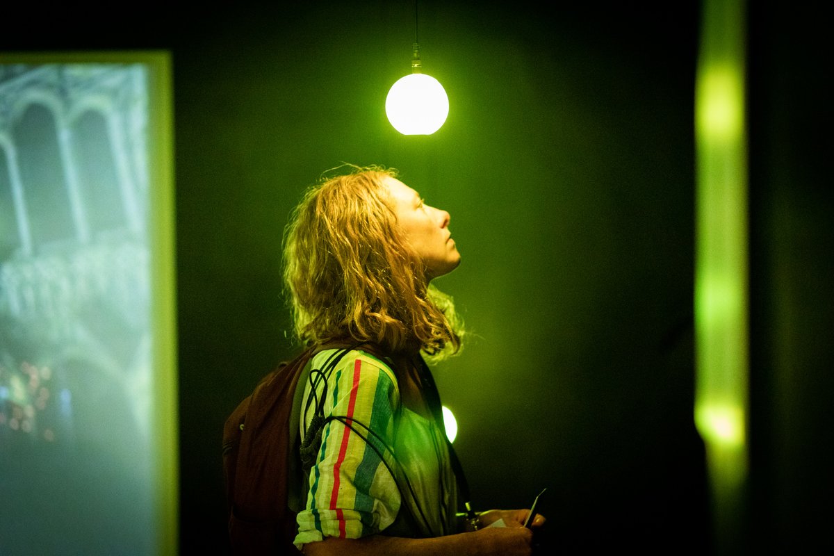 A woman with blonde hair and a stripy shirt stands under a green lightbulb. She looks off to the right of the picture.