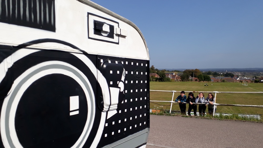 A group of young people stand next to a caravan painted as a camera.