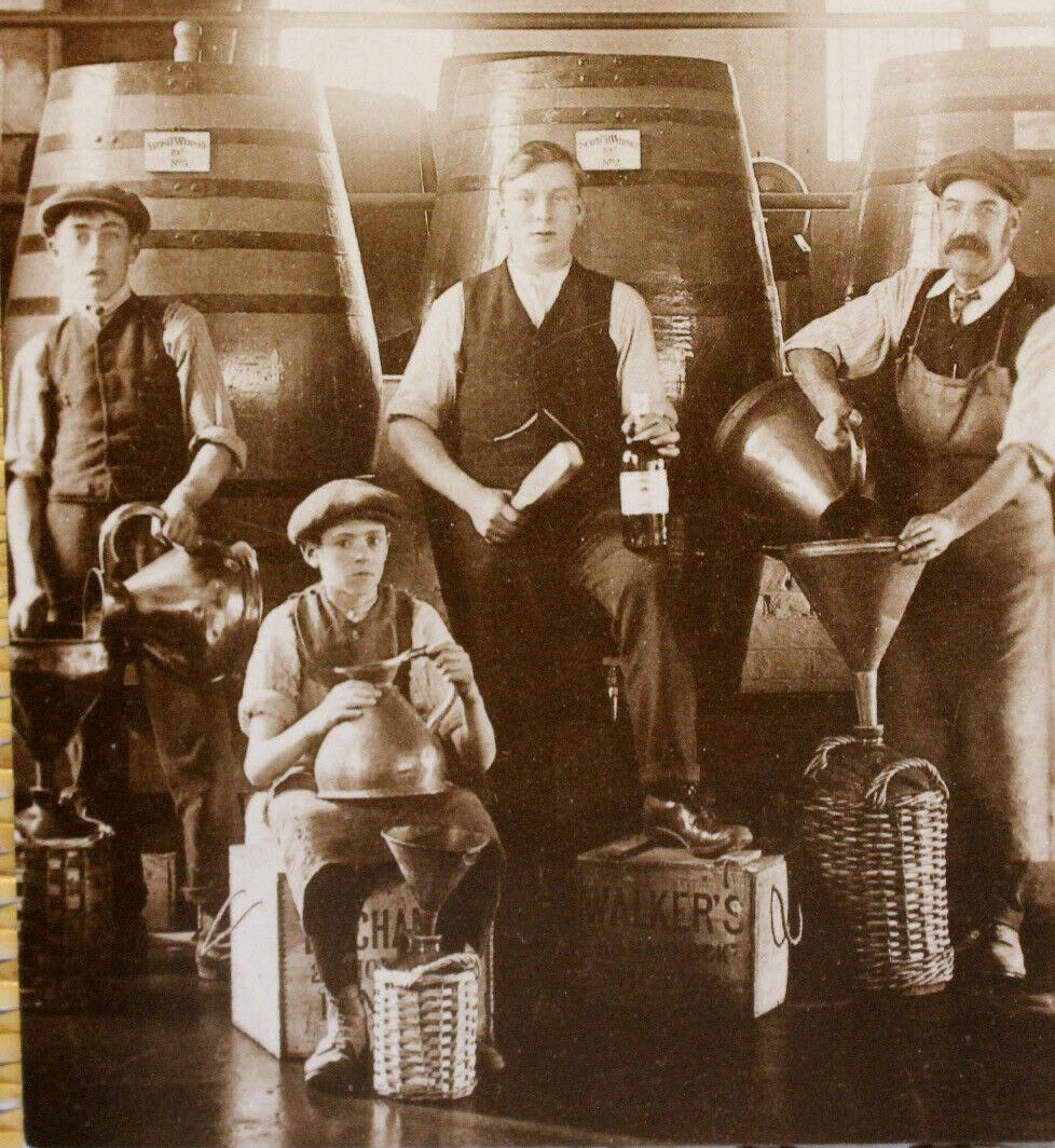 Early sepia photo of four men working at a whisky distillery, posing with their tools. Their ages range from around twelve to middle age.