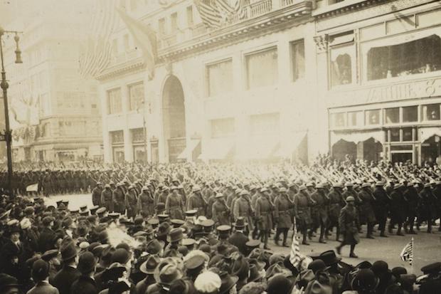 A black and white photograph shows the 369th Infantry Regiment, marching up Fifth Avenue during a parade with spectators.