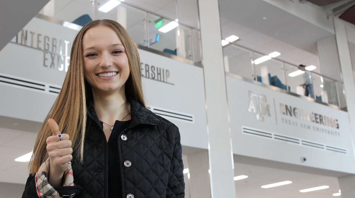 Ashley standing in an atrium, smiling, thumbs up gesture, natural light