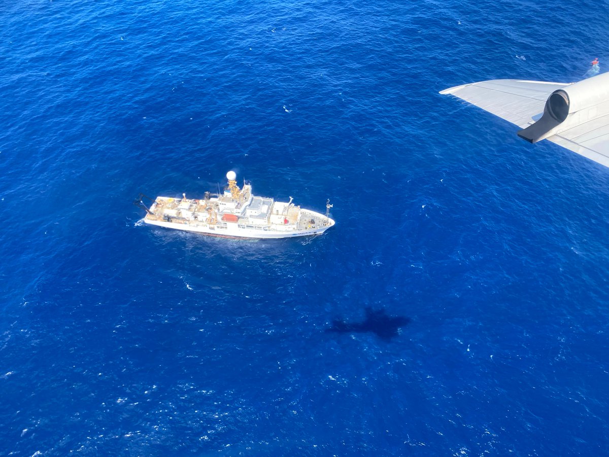 A large research ship is seen from above surrounded by blue water. The shadow of a plane is also seen on the water, and the plane's wing is visible in the top right corner. 