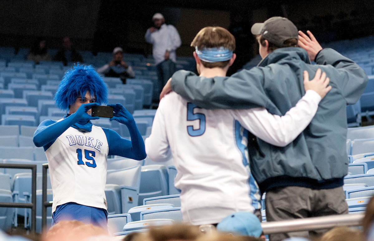 A man wearing a Duke basketball jersey takes a photo of two Carolina basketball fans