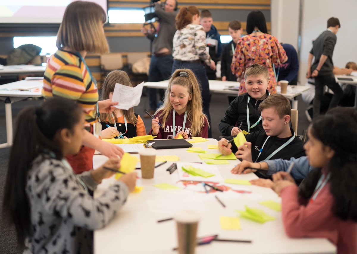 Group of children around a table, working together with a designer to post-it not ideas and create a physical prototype out of craft materials. 
