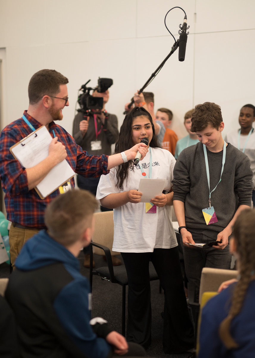 A young girl and boy presenting their project to a group. A microphone is being held to the girls mouth and a television camera and boom mic can bee seen in the background.