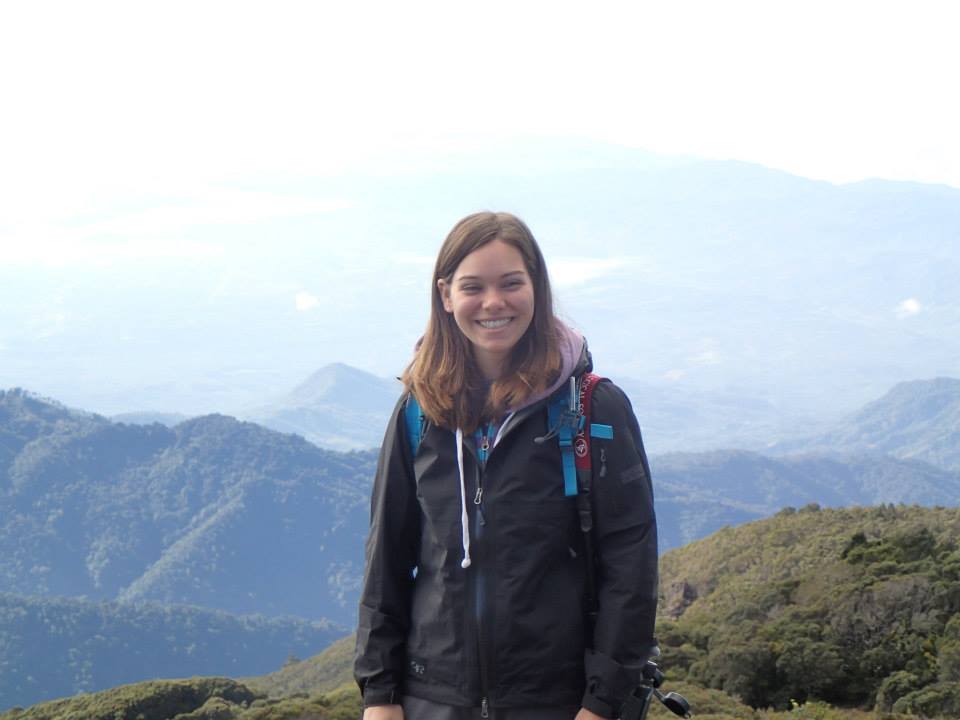 young woman dressed in hiking clothes, smiling, with a mountain view in the background