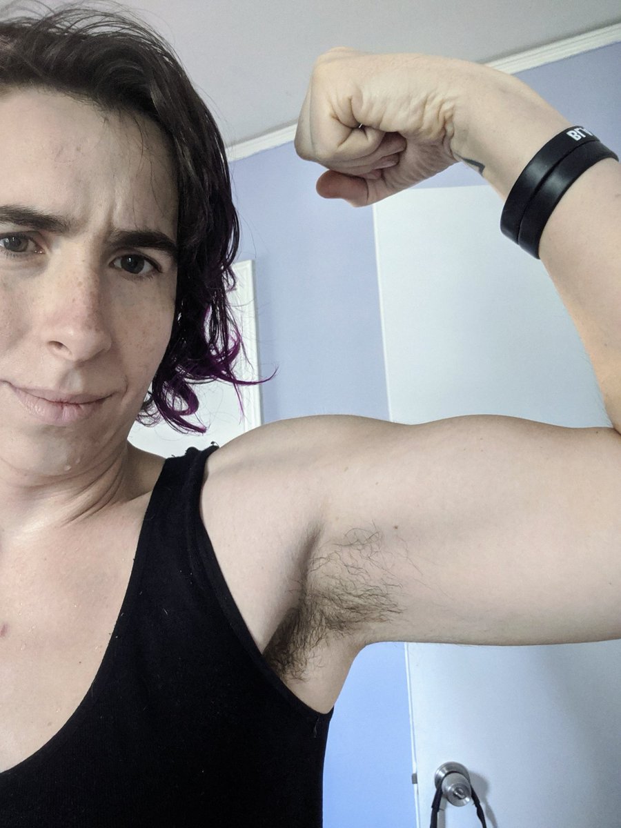 A dark-haired white trans woman in a black tank top flexing her right bicep. There is armpit hair present.